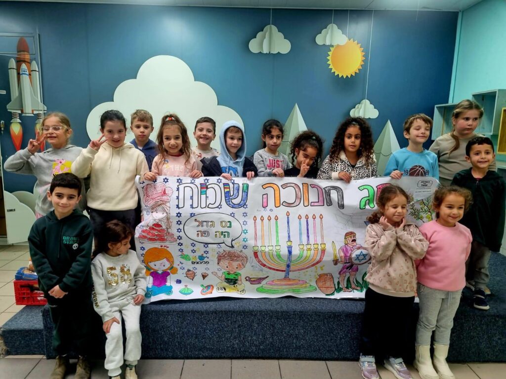 ילדים יוצרים סביב שולחן בקייטנה, צבעים ונרות חנוכה מפוזרים - אווירה שמחה וקהילתית