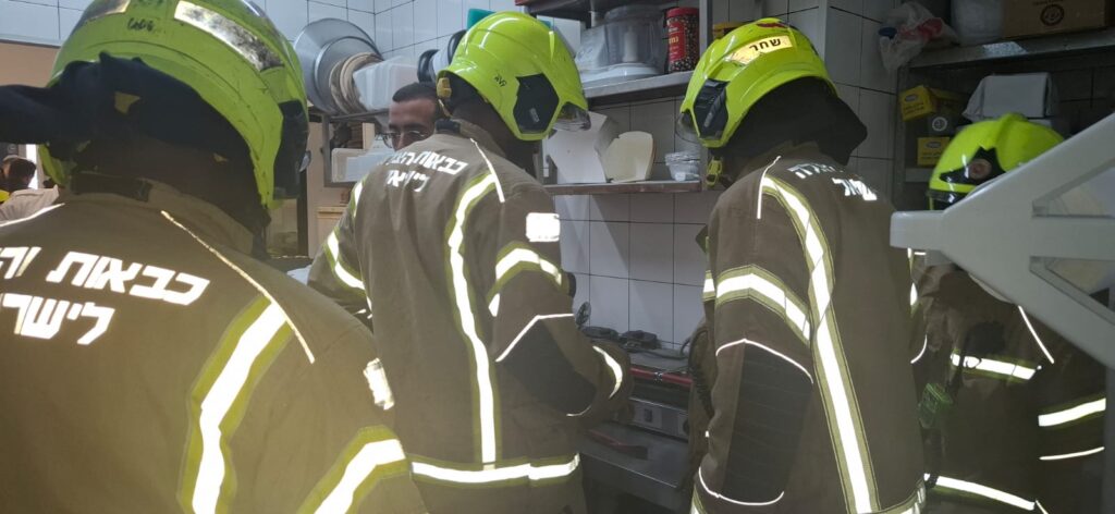 לוחמי אש עם ציוד חילוץ מקצועי מבצעים פירוק מדויק של מכשיר לישת בצק בתוך פיצרייה בקריית גת, כשצוות רפואי ממתין בסמוך