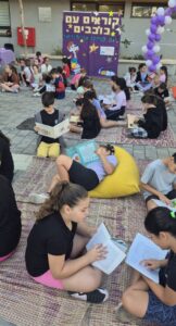 תלמידי כיתה א’ יושבים על מחצלות צבעוניות בחצר בית הספר, אוחזים ספרים ומתמסרים לקריאה. חיוכים, שקט טוב וקצת קסם של התחלה