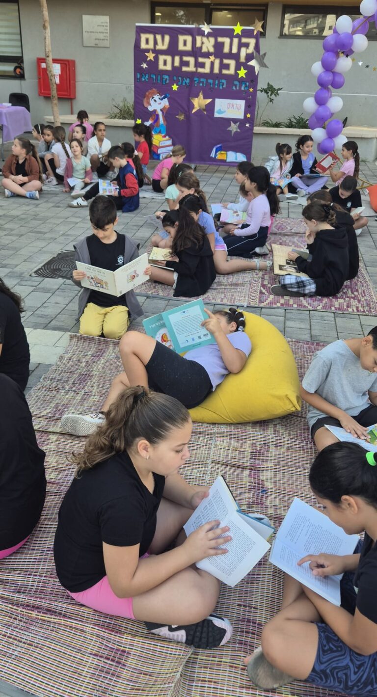 תלמידי כיתה א’ יושבים על מחצלות צבעוניות בחצר בית הספר, אוחזים ספרים ומתמסרים לקריאה. חיוכים, שקט טוב וקצת קסם של התחלה