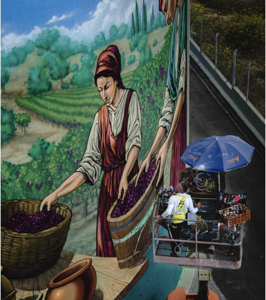 האמן על במת העבודה מול קיר ענק, מצייר דמות בוסקת ענבים. מאגר מים שהופך לציור קיר חי, צבעוני ומלא תנועה