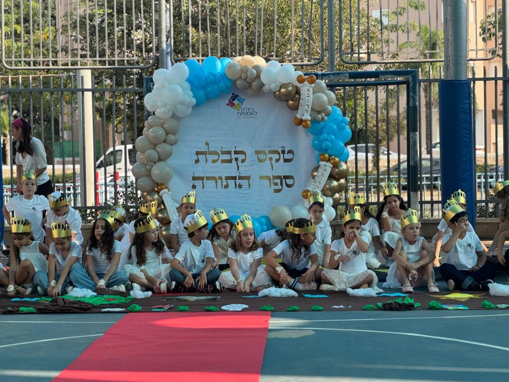 תלמידי כיתה ב’ מתהלכים עם ספר התורה לעבר בית הכנסת - הורים מלווים, דגלים, שירים וחיוכים. רגע של שמחה ואחדות.