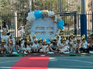 תלמידי כיתה ב’ מתהלכים עם ספר התורה לעבר בית הכנסת - הורים מלווים, דגלים, שירים וחיוכים. רגע של שמחה ואחדות.