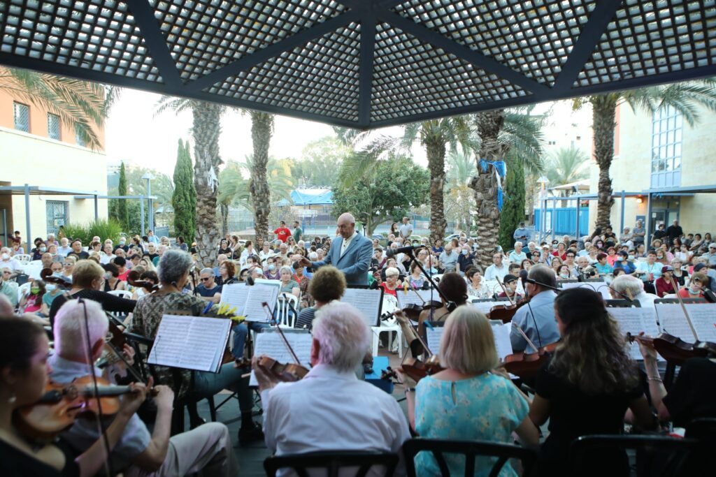 קונצרט קיץ פתוח בפארק שכונתי ברחובות, עם תזמורת מקומית וקהל משפחתי על מחצלות וכיסאות מתקפלים.