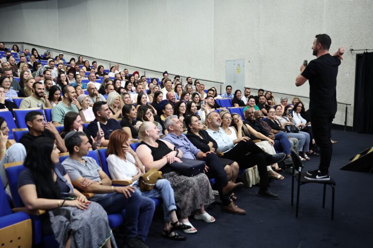 קהל גדול באולם תרבות ברמלה צופה במופע בידור חגיגי, כשברקע אווירה חמה של ערב הוקרה למשפחות המילואים והקבע