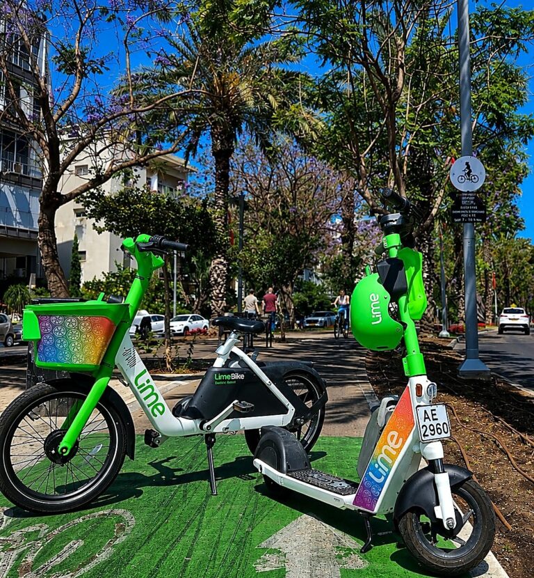 שורת קורקינטים ואופניים שיתופיים בצבעי הגאווה ניצבת לאורך טיילת תל אביב, כשברקע דגלי גאווה, רוכבים ועוברי אורח באווירת חגיגות הקיץ בעיר