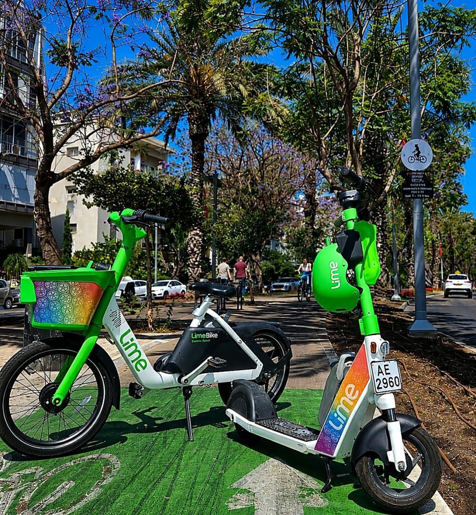 שורת קורקינטים ואופניים שיתופיים בצבעי הגאווה ניצבת לאורך טיילת תל אביב, כשברקע דגלי גאווה, רוכבים ועוברי אורח באווירת חגיגות הקיץ בעיר
