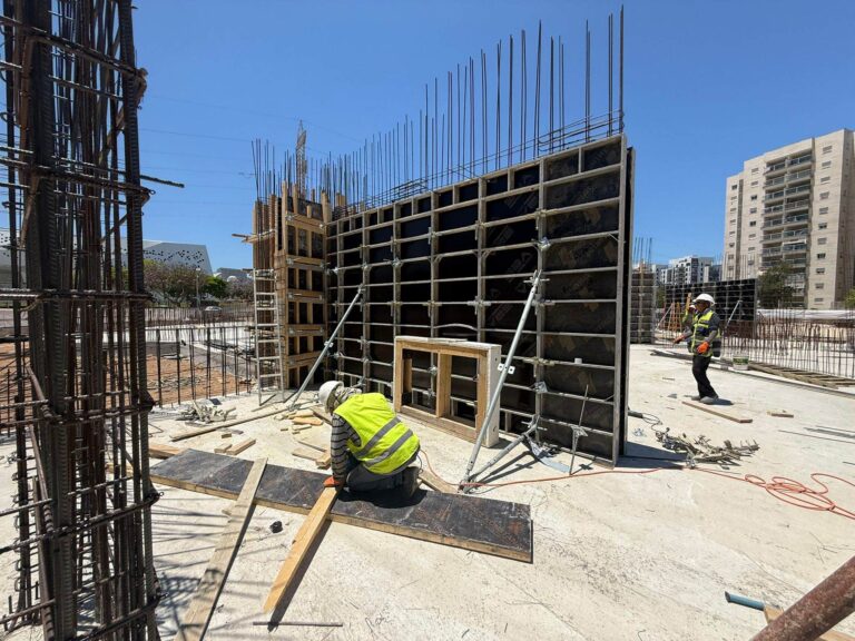 אתר בנייה מודרני באשקלון עם עבודות יסוד מתקדמות, כשברקע הדמיה אדריכלית של מרכז צעירים חדש הכולל מרחבים קהילתיים, כיתות לימוד ואודיטוריום