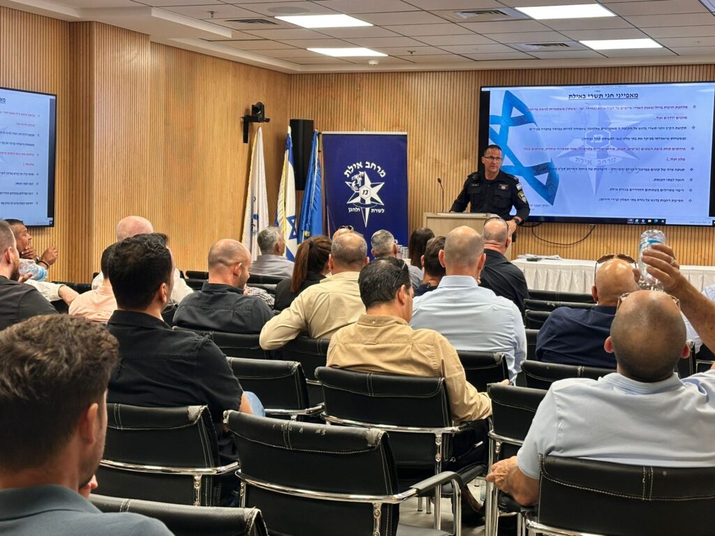 שוטרי מרחב אילת ובעלי עסקים יושבים בכנס היערכות לקיץ באולם עירוני, כשברקע מצגת מקצועית על ביטחון, תיירות ומוקדי בילוי בעיר