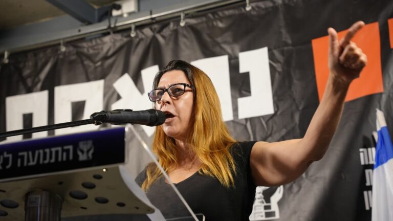עו"ד גייל שורש נואמת על במה מול קהל בכיכר הבימה, כשברקע דגלי ישראל ומשתתפי הפגנה, באווירה ציבורית וערכית