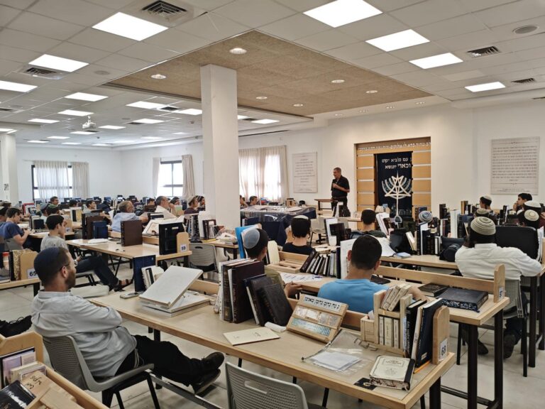 האלוף במיל' יפתח רון טל משוחח עם תלמידים באולם ישיבה תיכונית, כשברקע קהל תלמידים מאזין להרצאה העוסקת במנהיגות, ערכים ושירות ציבורי.