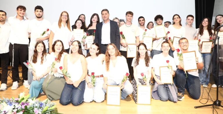 בני ובנות נוער ממודיעין מכבים רעות עומדים על במה בטקס הוקרה עירוני, מחזיקים תעודות הצטיינות לצד נציגי העירייה וקהל משפחות באולם