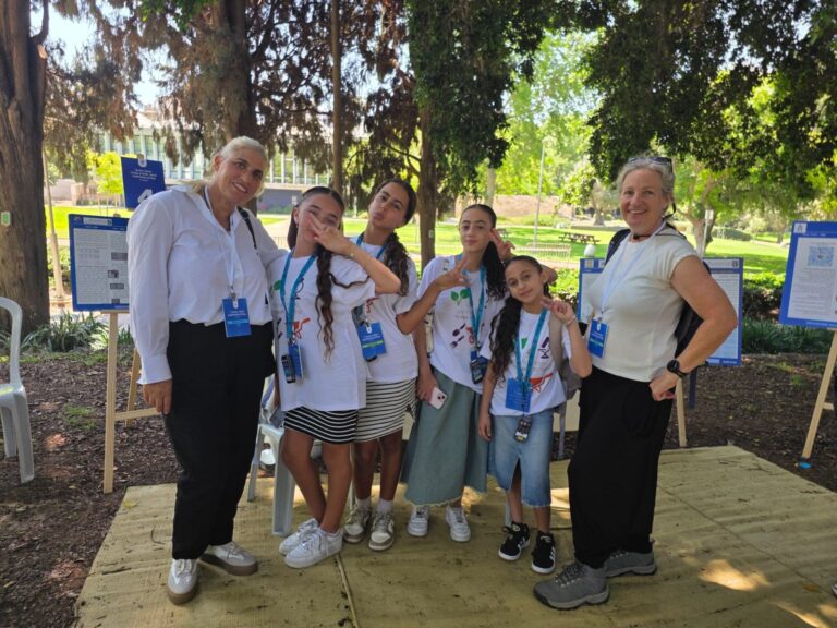 תלמידות בית ספר עומדות לצד מתקן גידול נבטים בחווה חקלאית, כאשר ברווזים אוכלים צמחייה ירוקה מתוך המתקן שפיתחו במסגרת פרויקט מדעי.