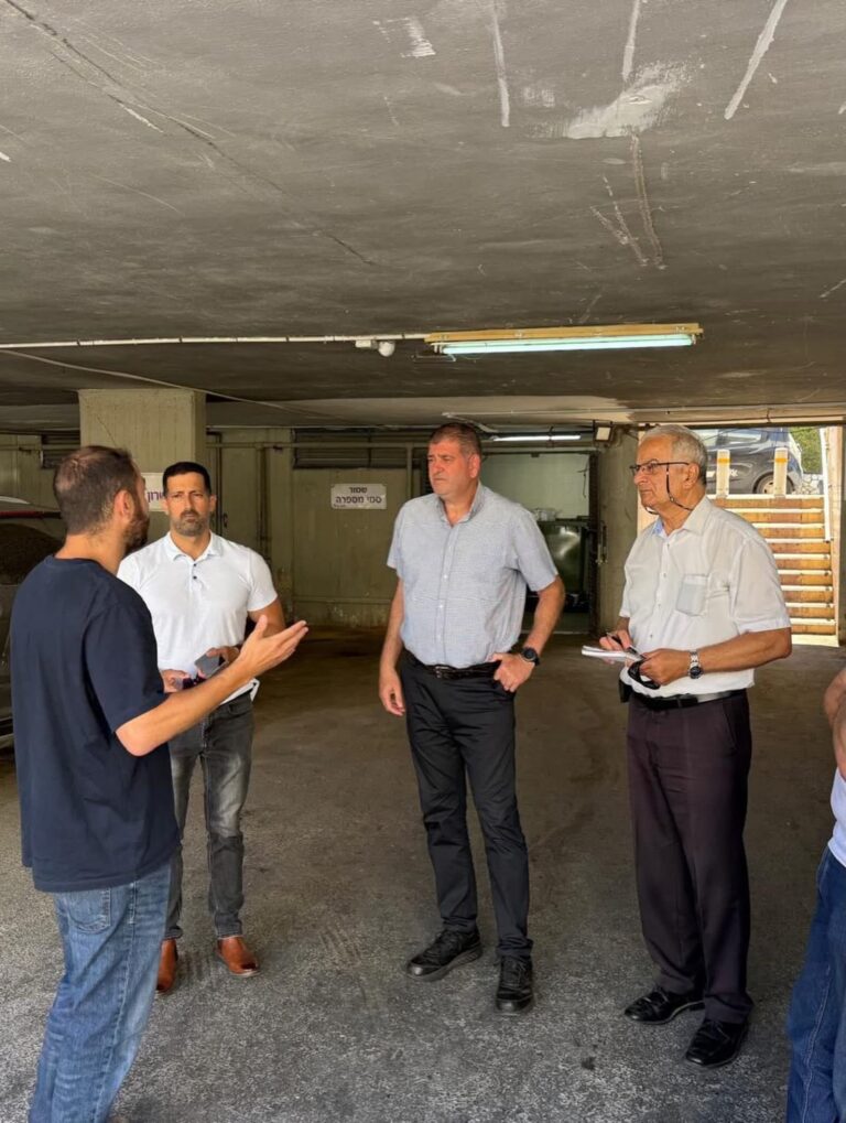 מרכז מסחרי שכונתי בגבעת שמואל במהלך עבודות ניקיון ושדרוג, עם בעלי עסקים, תושבים, פחי מחזור, דחסן קרטונים וחזיתות חנויות באזור ציבורי מטופח
