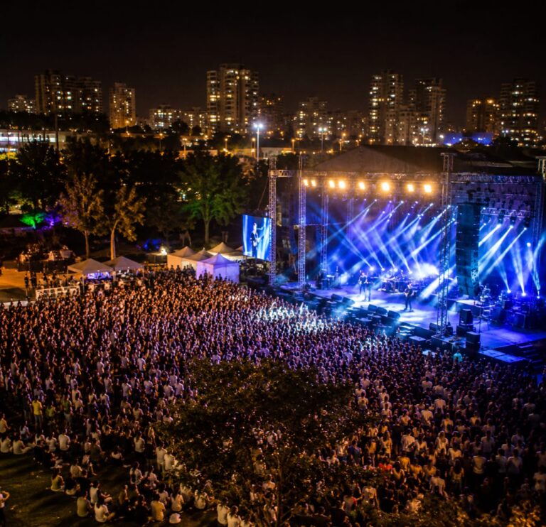 פארק עירוני חדש בראשון לציון בשעת ערב, עם במה מרכזית, קהל רב, תאורת מופע, מדשאות רחבות ואווירת חגיגת קיץ עירונית