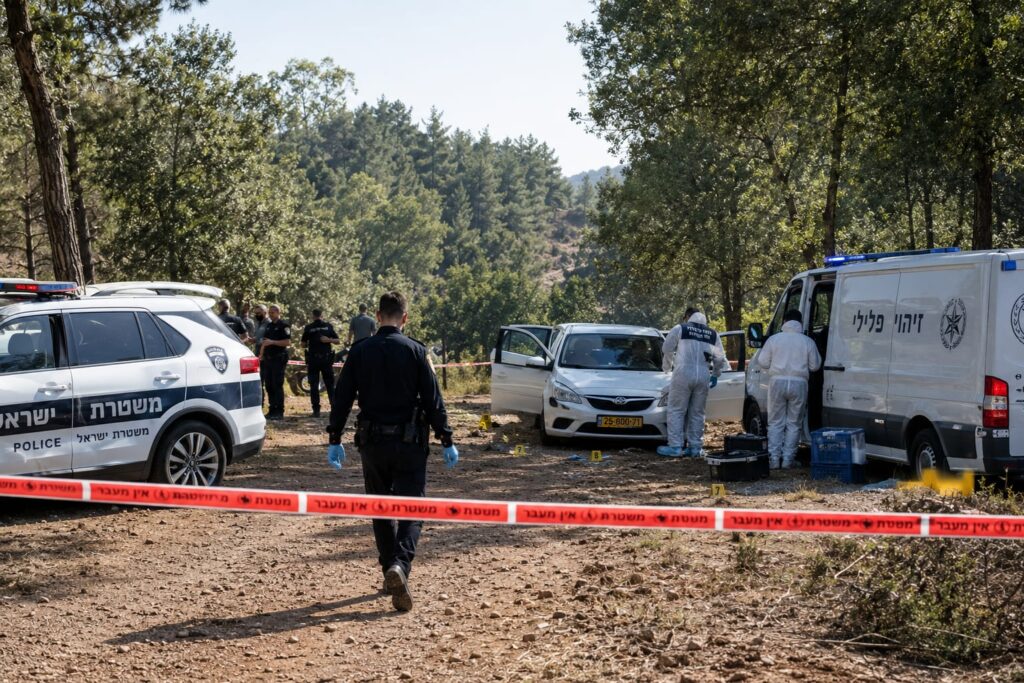 ניידות משטרה וחוקרי מז"פ בזירת יער סמוכה למעיין ורדה, לצד רכב פרטי, סרטי סימון וציוד חקירה, באווירה של זירת רצח חמורה