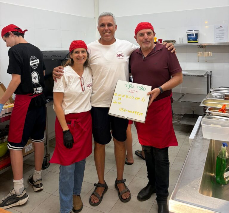 בני נוער עובדים יחד בפיצרייה קהילתית בקיבוץ, מכינים פיצות ומגישים ללקוחות, כשברקע אווירה חמה של קהילה, התנדבות ושיתוף פעולה בין צעירים לתושבים