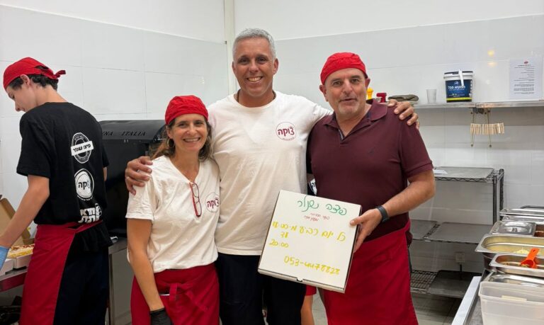 בני נוער עובדים יחד בפיצרייה קהילתית בקיבוץ, מכינים פיצות ומגישים ללקוחות, כשברקע אווירה חמה של קהילה, התנדבות ושיתוף פעולה בין צעירים לתושבים
