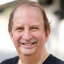 סולי תמרי, יזם בן 75 מהרצליה, יושב מול מחשב ומדגים מערכת ללימוד אנגלית, כשברקע תלמידים מתרגלים שיחה באנגלית באמצעות טכנולוגיה מתקדמת, באווירה של חדשנות וחינוך