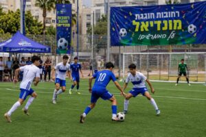ילדים, בני נוער ומבוגרים משתתפים בטורניר כדורגל קהילתי במגרש ספורט ברחובות באווירת מונדיאל