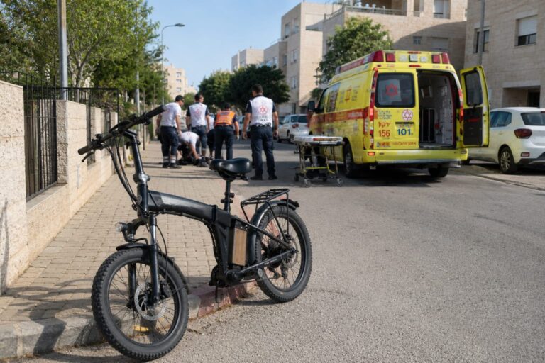 אופניים חשמליים מונחים בצד הדרך ברחוב מגורים בלהבים, כשברקע ניידת טיפול נמרץ וצוותי הצלה