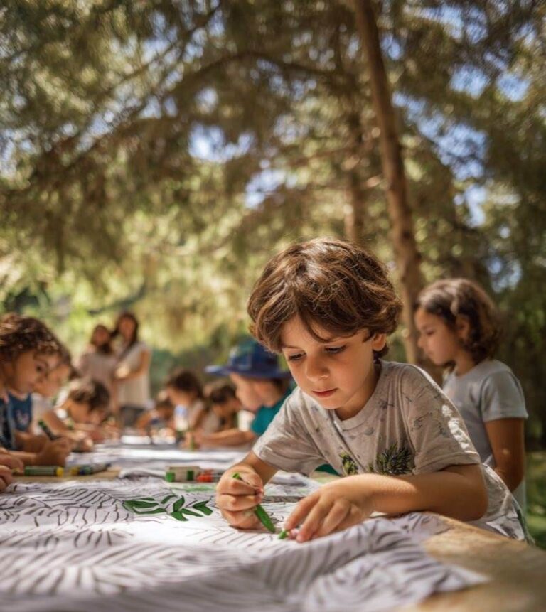 ילדים ומשפחות משתתפים בפעילות חווייתית באירוע קיץ בפארק או בחצר בית ספר באשקלון, עם מתחמי יצירה, משחקים והצגה ברקע
