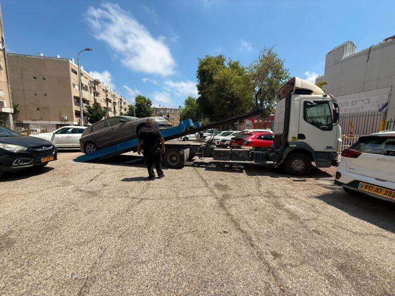 גרר עירוני מפנה רכב ישן ונטוש מרחוב מגורים באשדוד, לצד פקחים עירוניים, מדרכה נקייה ובנייני מגורים ברקע