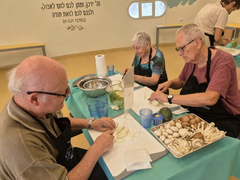 אזרחים ותיקים מבשלים יחד במטבח קהילתי מודרני בשכונה ט' בבאר שבע, כשברקע מבנה ציבורי משופץ, באווירה חמה ומחברת