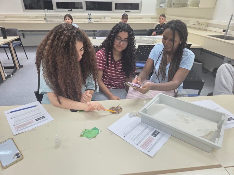 קבוצת תלמידים ותלמידות במעבדת מדע מתקדמת במכון ויצמן, לצד רובוט לימודי ומסכי מחשב, באווירת חדשנות ומצוינות