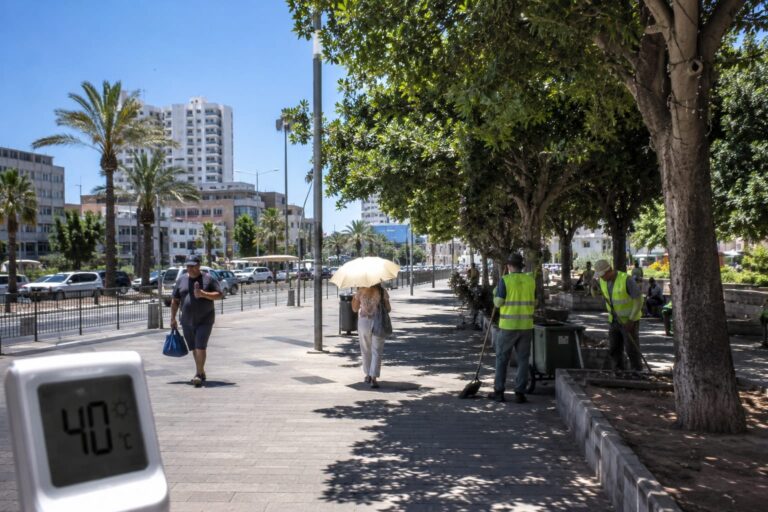 רחוב מרכזי באשדוד בשעות הצהריים תחת שמש קופחת, מד חום המראה טמפרטורה גבוהה, מעט הולכי רגל המחפשים צל ועובדי עירייה מבצעים עבודות באזור מוצל